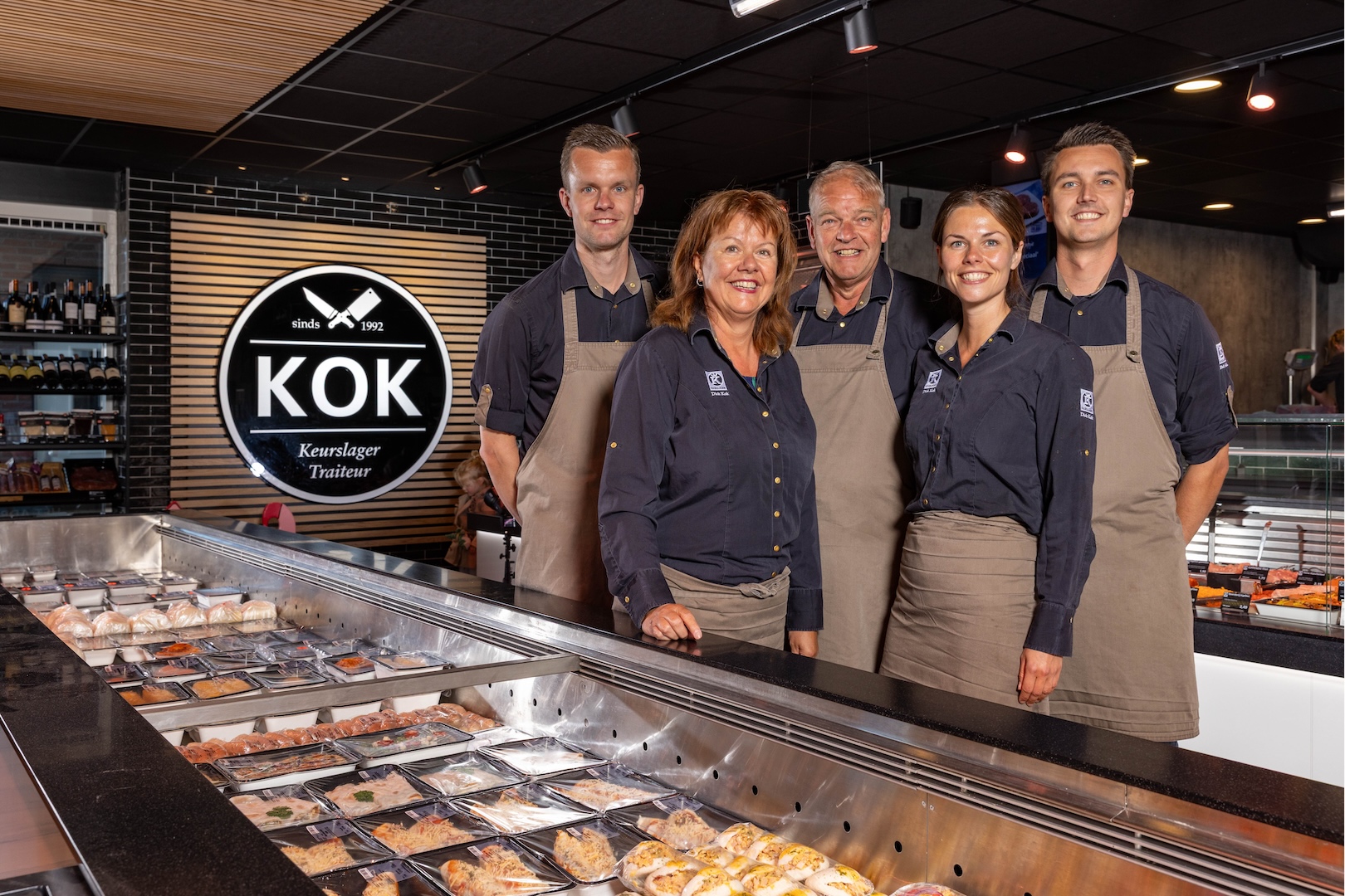 Het team van Keurslager en traiteur Kok voor de toonbank, met het eigen KOK-logo op de achtergrond